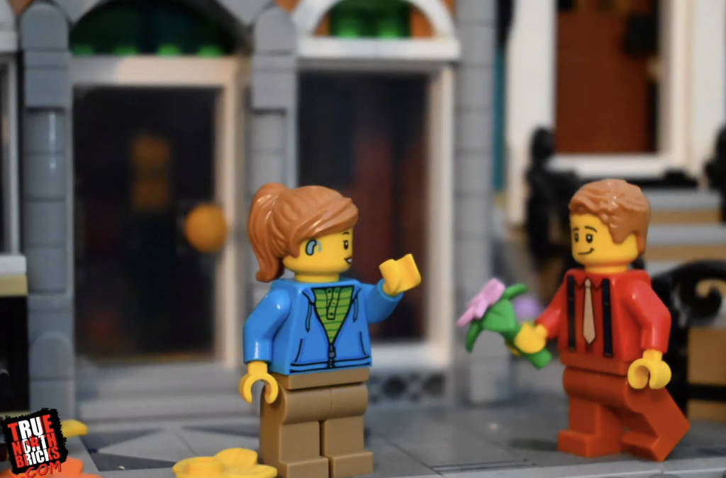 Two Lego figures stand in their lego world. The figure on the left has a ponytail and a hearing aid. She is wearing a blue hoodie and brown pants. The figure on the right is wearing a red suit and has flowers in his hands. Lego Diversity with Sign Language Blitz.