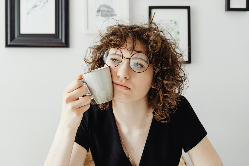Woman sits in thought. She has curly hair and is wearing glasses. She is holding a coffee cup against her face.