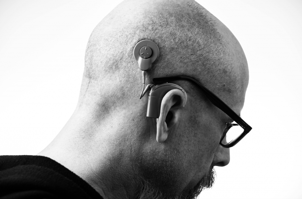 A man looks down and to the right. He is bald, wearing black glasses, and a hearing aid.